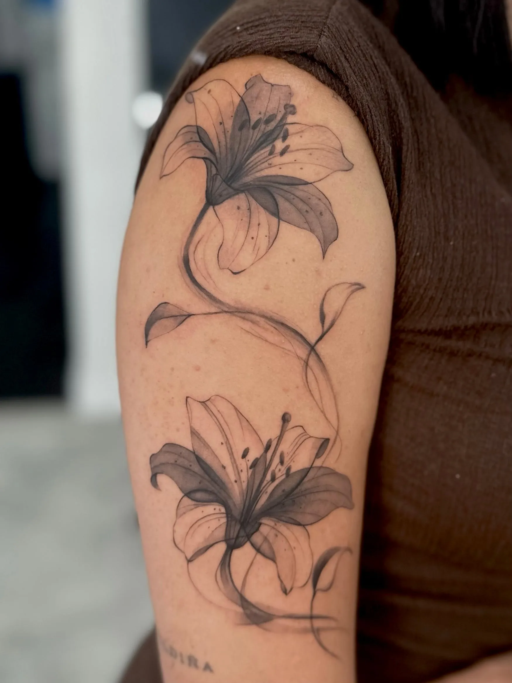 Dos azucenas en negro y gris con efecto translúcido, unidas por un tallo ondulante, en el brazo