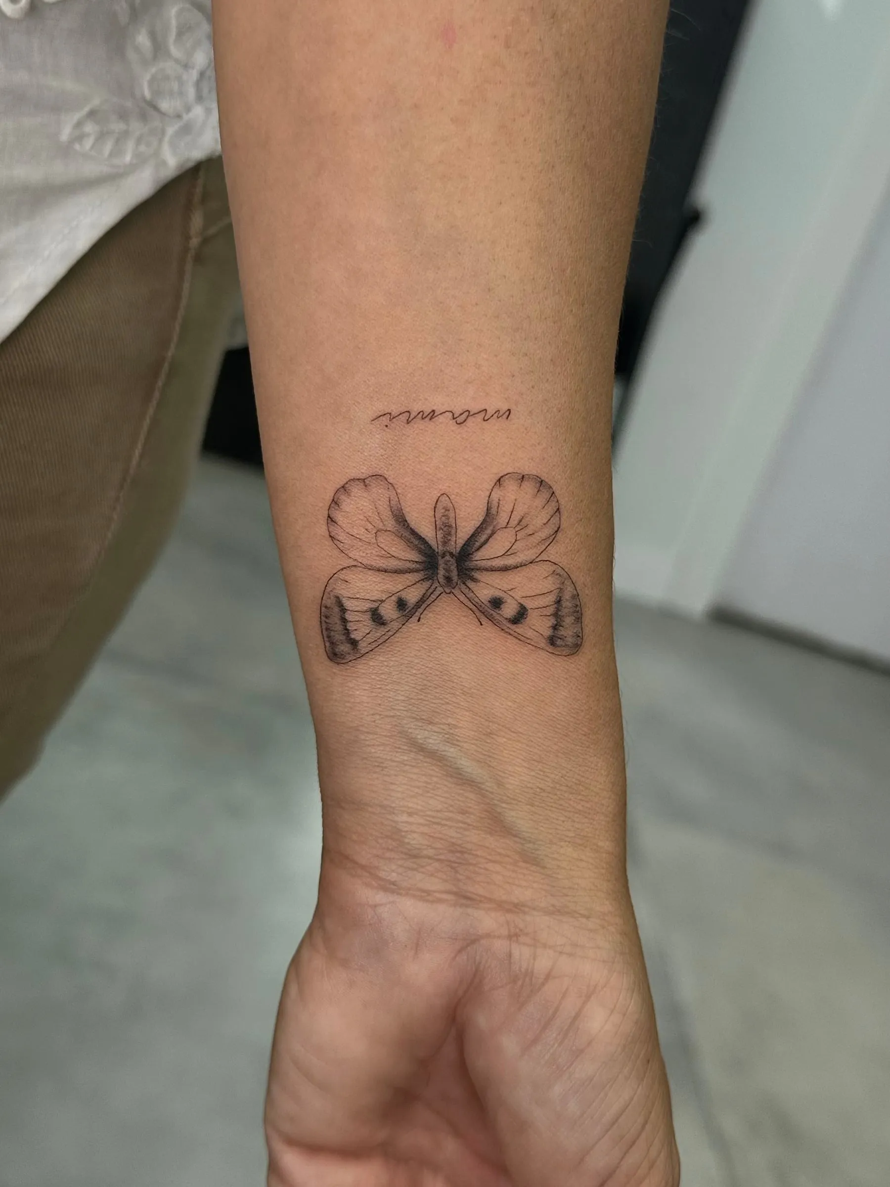 Mariposa en línea fina con sombreado suave y una pequeña palabra manuscrita encima, sobre la muñeca