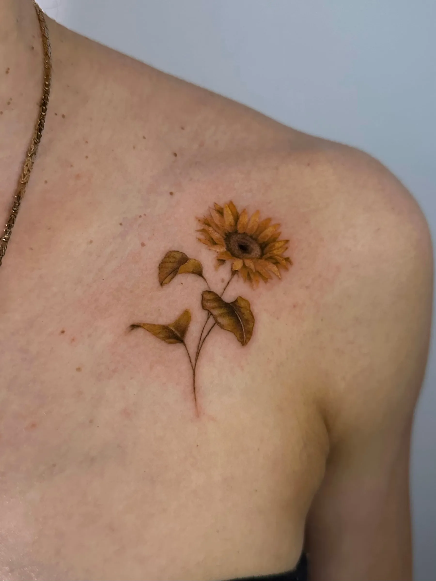 Girasol a color en tonos dorados con hojas, bajo la clavícula