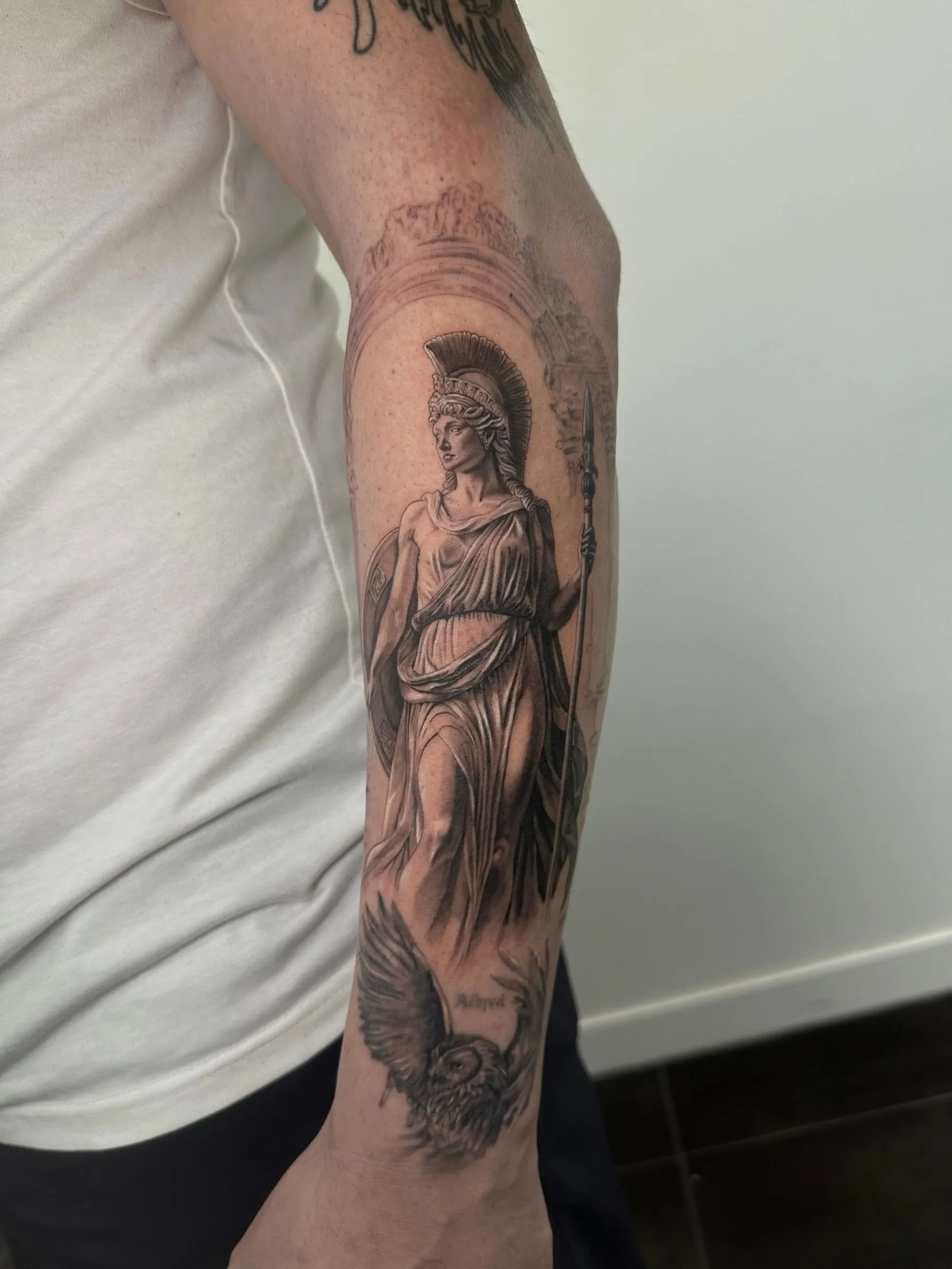 Figura realista en negro y gris de una diosa griega con casco y lanza, con un búho a sus pies, a lo largo del antebrazo